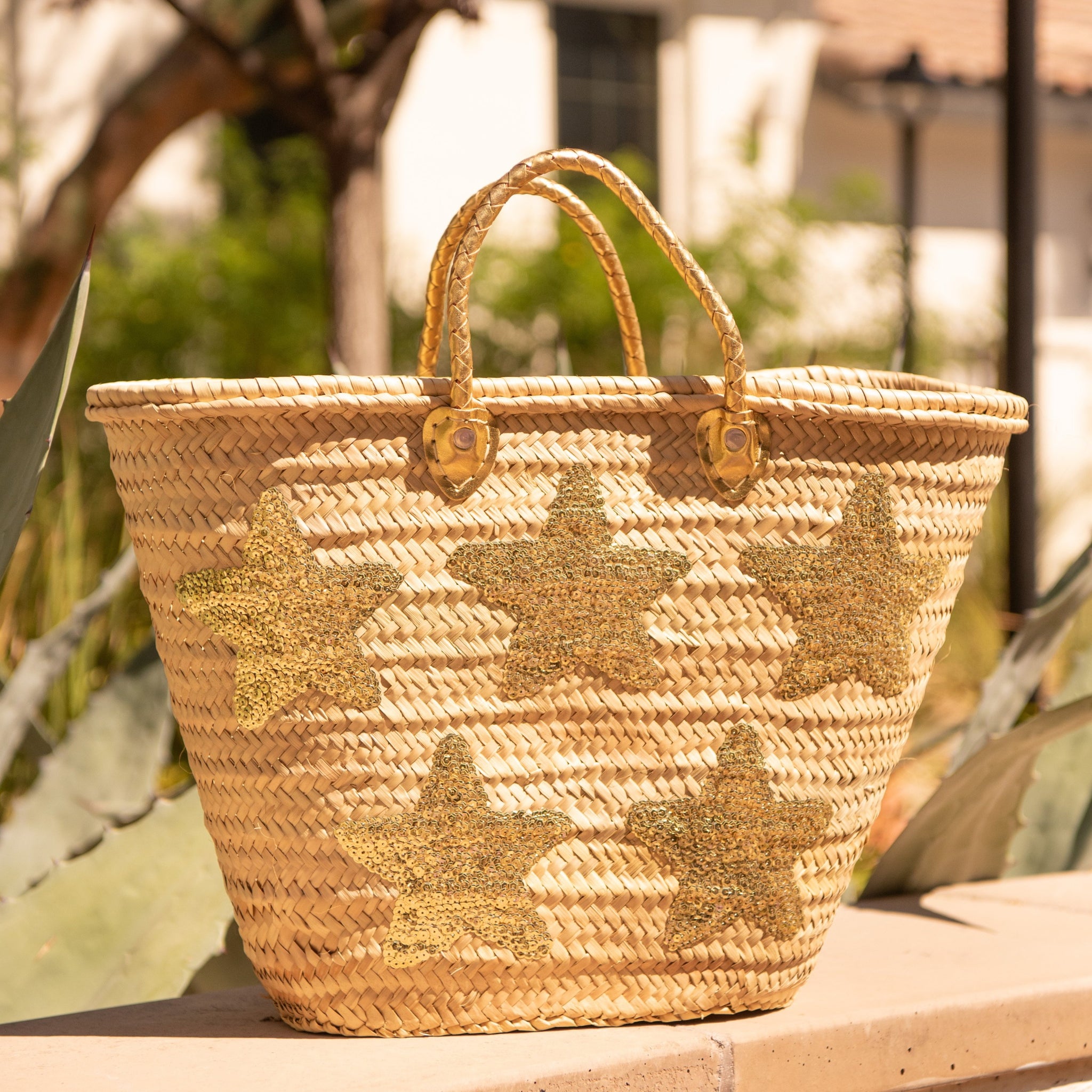 Large Straw Bag with Gold Sequin Stars sitting on ledge with trees behind it