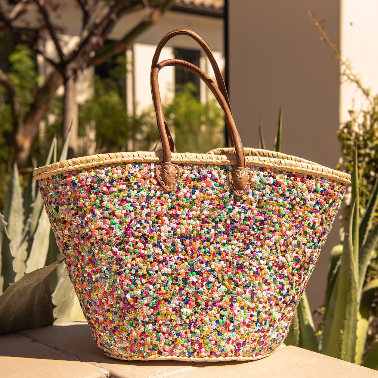 Large Straw Shoulder Bag with Multi-Colored Sequins sitting on ledge with green cactus behind 